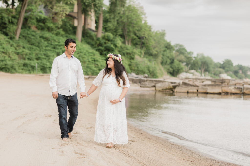 husband and wife holding hands walking on beach for maternity photoshoot 