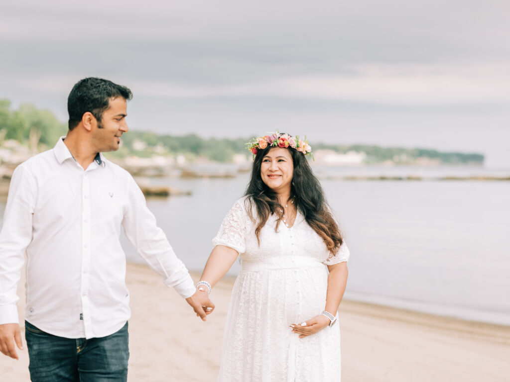 husband and wife holding hands walking on beach for maternity photoshoot 