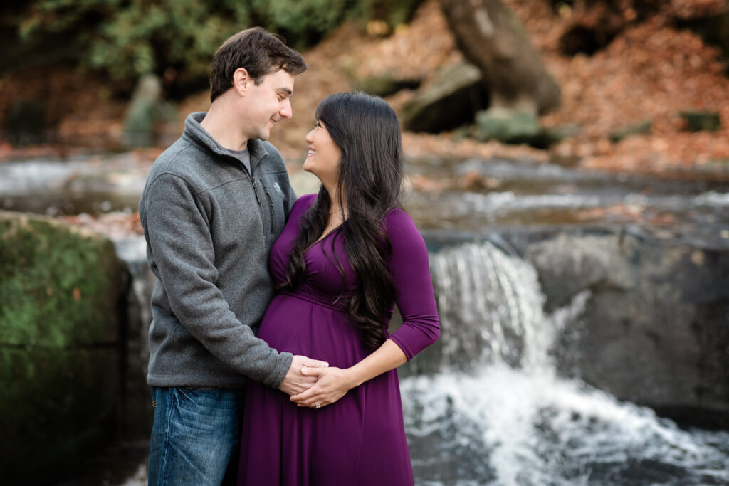 husband and wife holding pregnant belly by waterfall for maternity photoshoot The Womb Wellness Center 