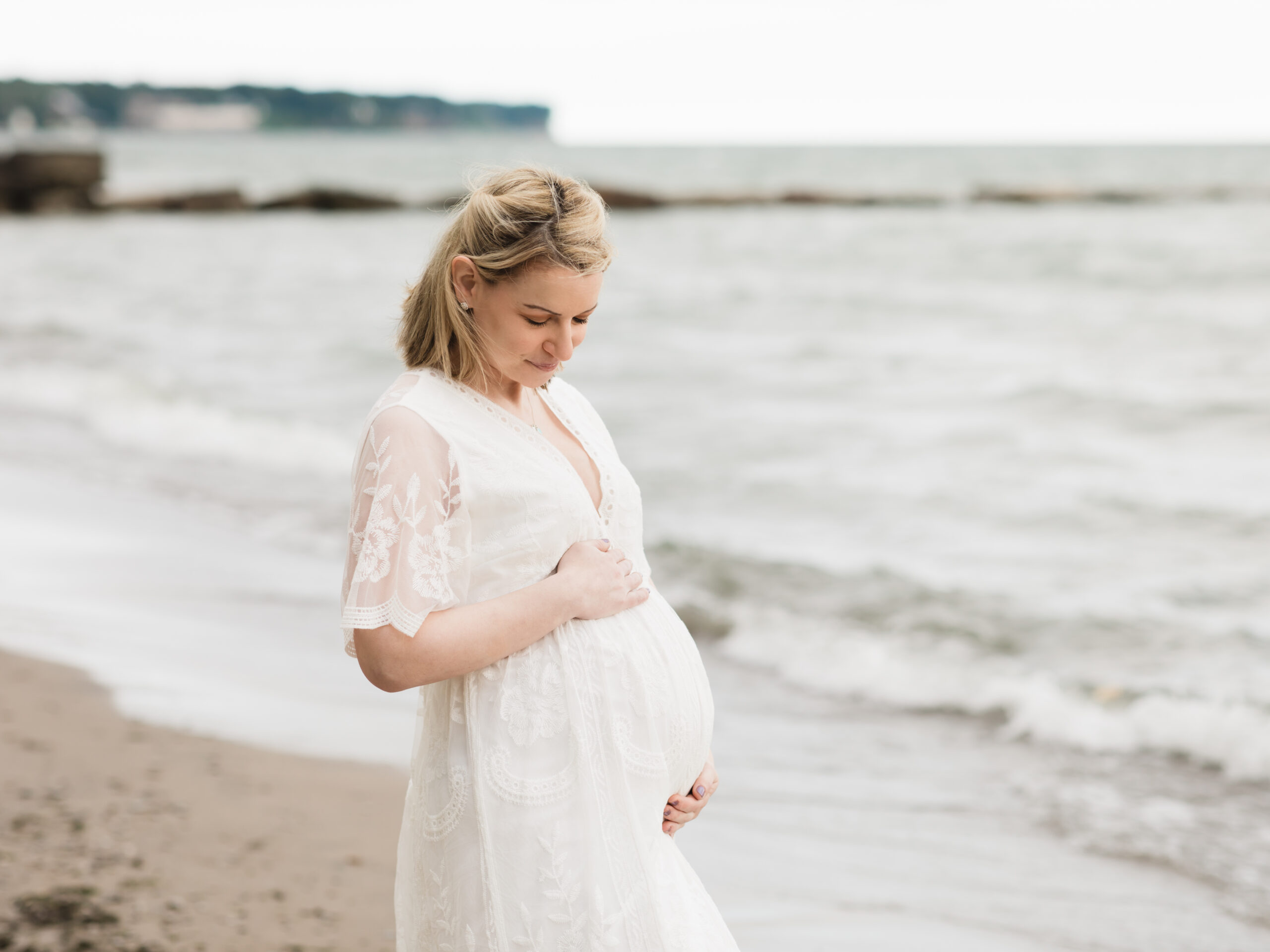 pregnant mother in white lace dress holding belly at the beach Fairview Hospital Birthing Center