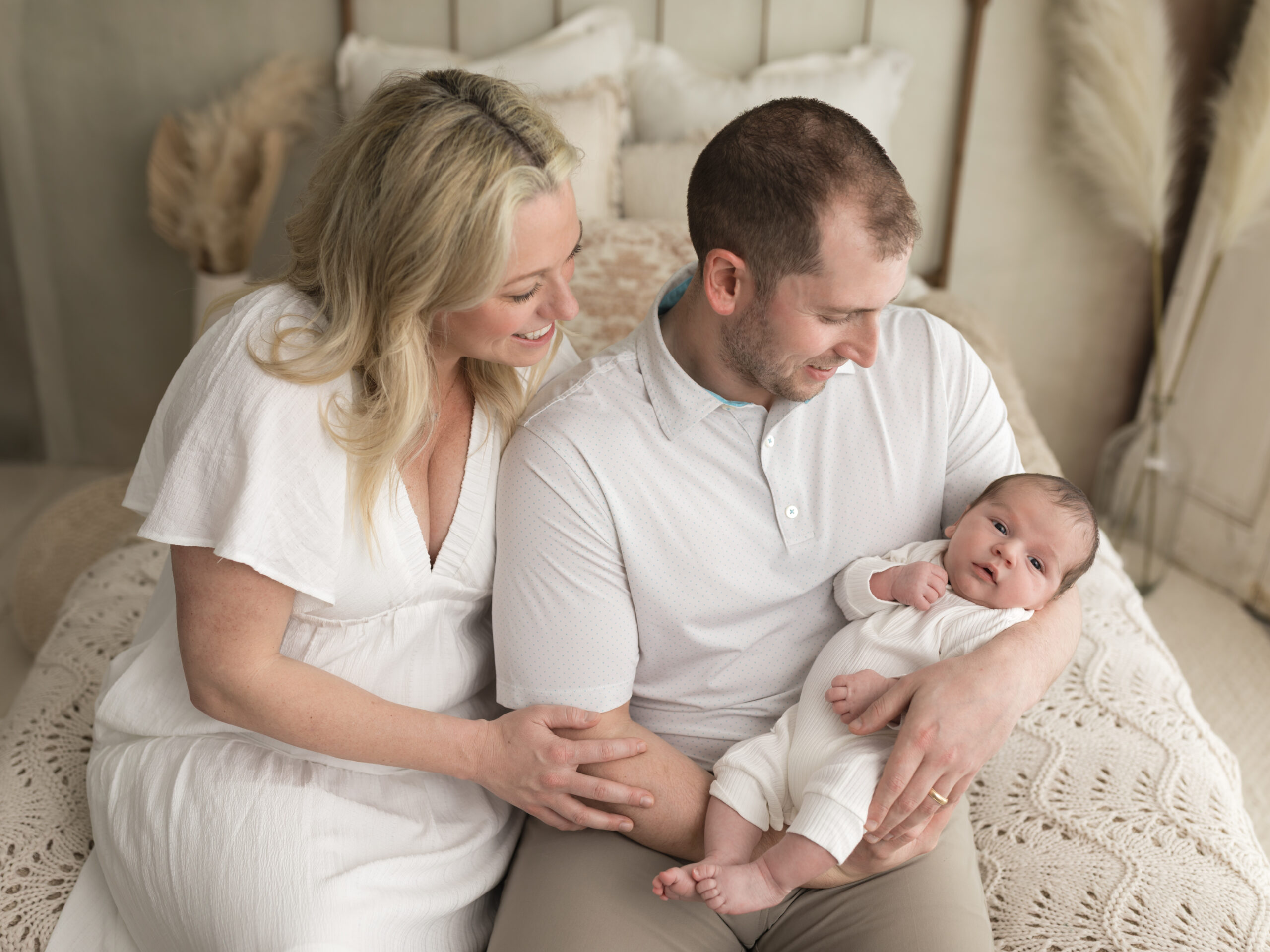 mom and dad dress in white sitting on bed holding newborn baby boy for studio portraits Akron Ohio Newborn Photographer