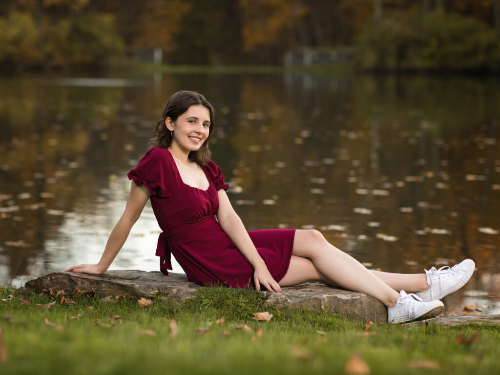 female senior in red dress sitting by pond for portraits