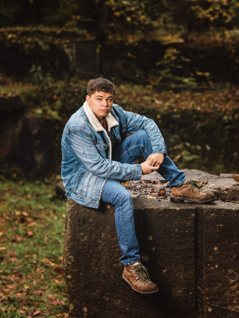 senior boy in jean jacket sitting on rock at camp manatoc for senior portraits