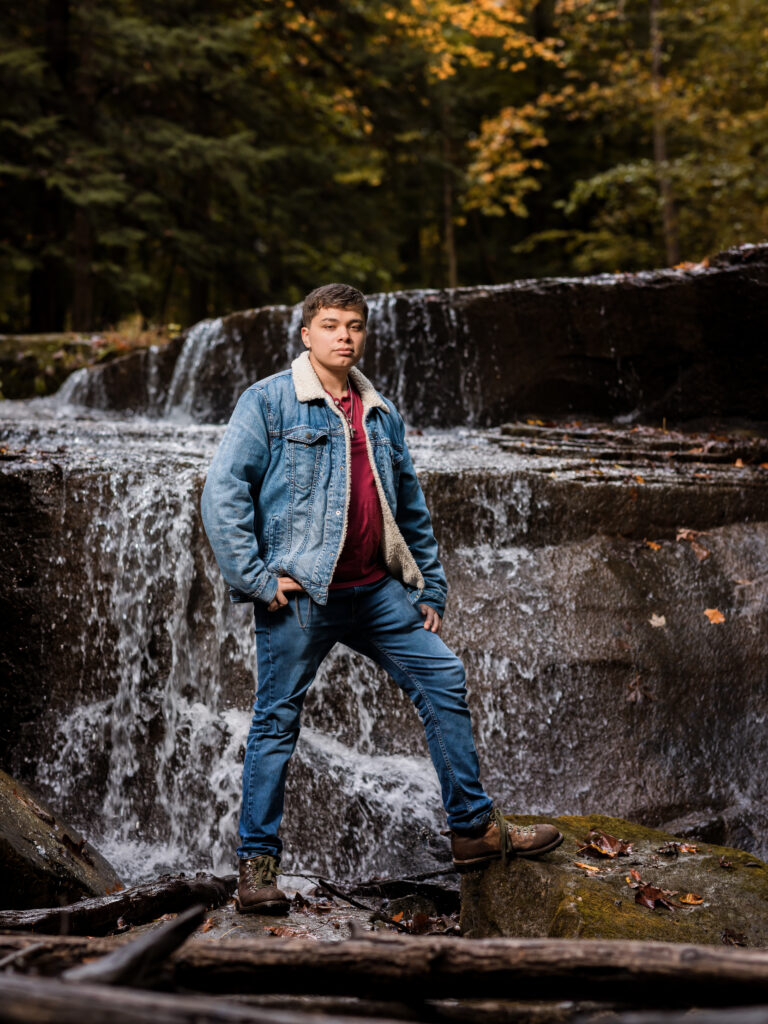 senior boy in jean jacket posed by waterfall for senior portraits