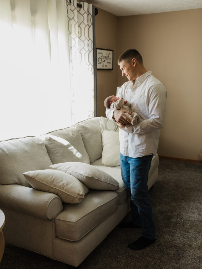 dad standing in living room holding newborn baby boy for in home newborn photoshoot Hatch Boutique