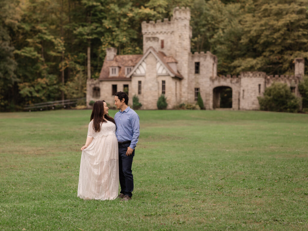 husband and pregnant wife in white lace dress standing in front of squires castle