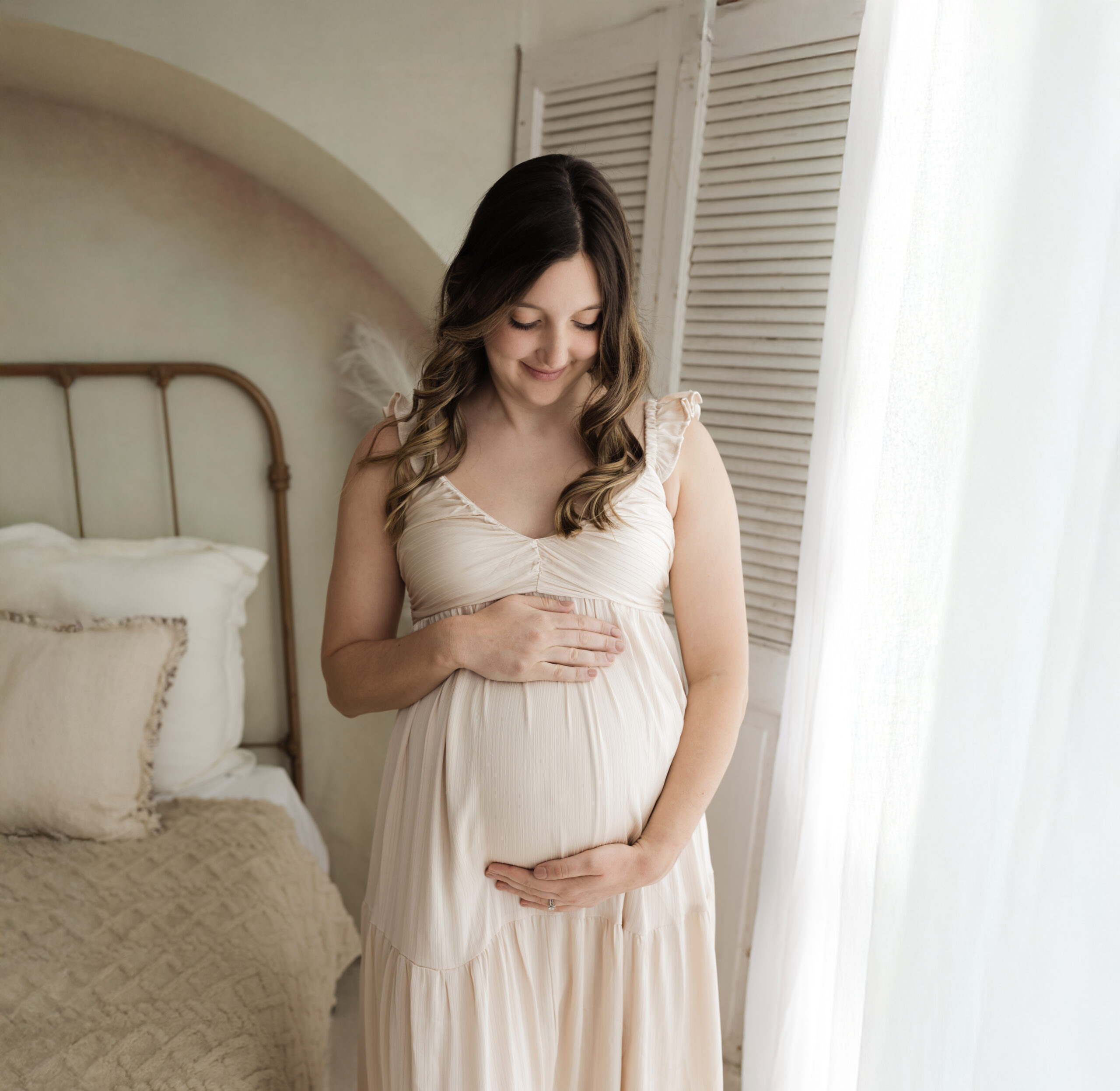 pregnant mother in cream maternity gown holding baby bump by window for maternity photoshoot Cleveland OBGYN