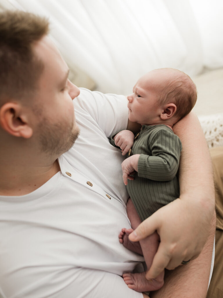 dad holding newborn baby boy in green onesie next to window 