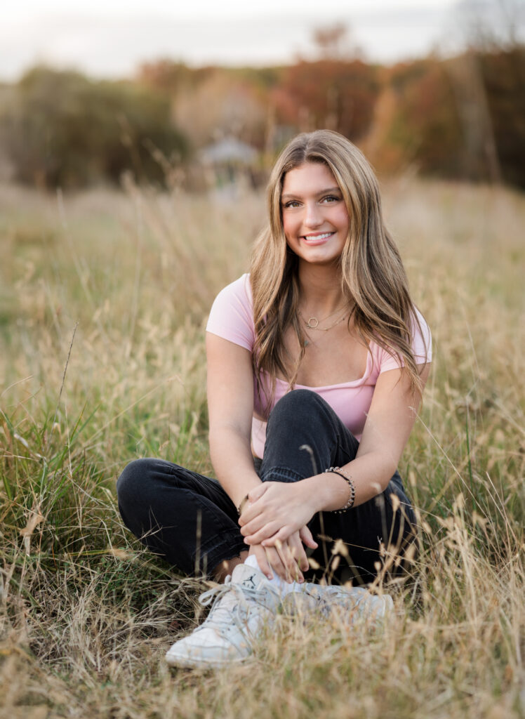 high school senior girl in pink shirt sitting in field at park for senior portraits 