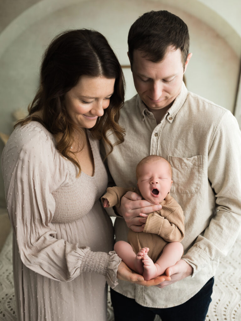 mom and dad holding newborn baby boy in tan onesie for studio newborn photos
