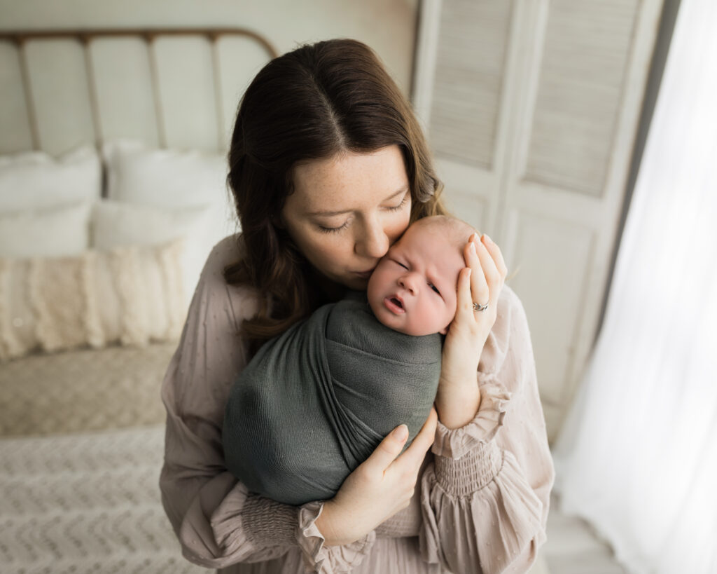 mom kissing newborn baby boy swaddled in green wrap Newborn Photography Guide